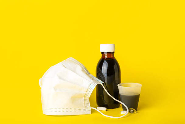 Бутылка сиропа от кашля, чашка, таблетки и медицинская маска на желтом фоне - Фото, изображение