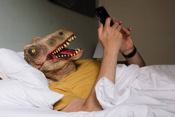 Άνθρωπος με μάσκα σαύρας χρησιμοποιώντας το smartphone σε ένα κρεβάτι ξενοδοχείου. - Φωτογραφία, εικόνα