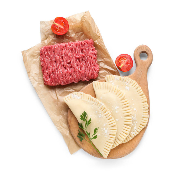 Board with raw chebureks, baking paper, mince and cut tomato on white background - Photo, Image