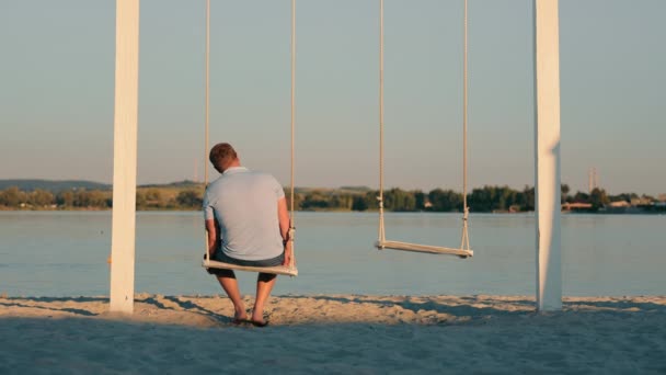 Achteraanzicht van een eenzame man die alleen zwaait op een schommel op het strand. Mannelijke eenzaamheid aan de oever van het meer - Video