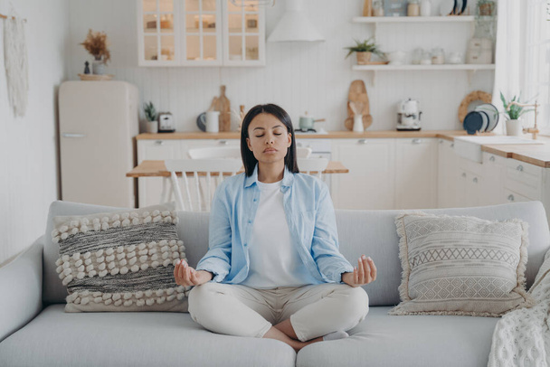 Serene woman practicing yoga and meditation keeping hands in mudra gesture, sitting cozy couch at home. Calm young female relaxing on sofa. Wellness, stress relief, emotion management. - Φωτογραφία, εικόνα