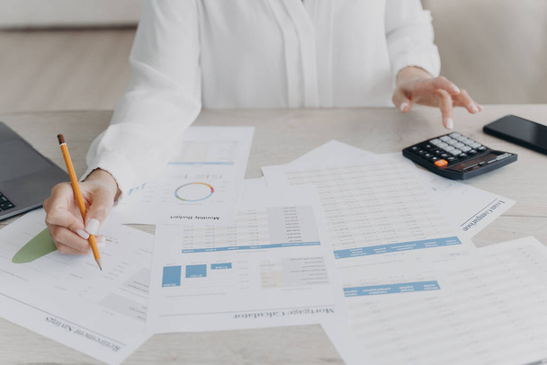 Close-up businesswoman calculating expenses, managing project budget, sitting at desk with documents. Woman counts finances, preparing report in charts and graphs, analyzing financial statistics. - Foto, Imagen