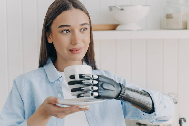 Confident handicapped girl is holding cup of tea with cyber hand. Happy disabled girl at kitchen at home. Pretty woman with makeup is smiling. Concept of grasp sensors in modern electronic prosthesis. - Photo, Image