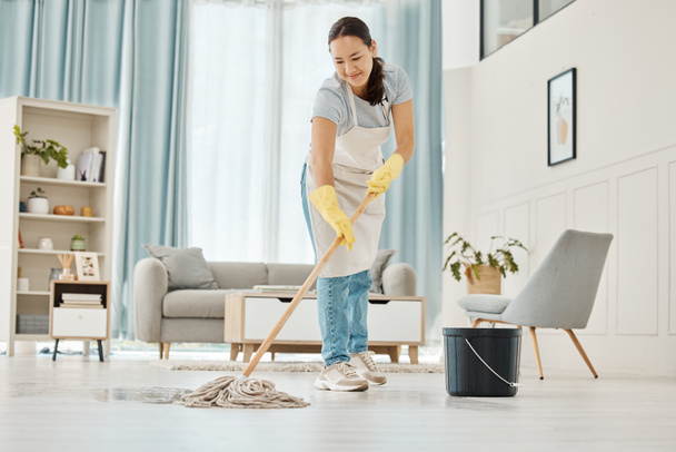 Egy nő, aki mosollyal takarítja a padlót a nappaliban. Boldog ázsiai takarító csinál házimunkát vagy munkát egy tiszta társalgóban, szállodai szobában vagy házban, miközben mosolyog és egyedül tavaszi takarítás. - Fotó, kép