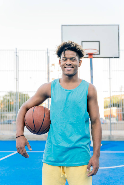 Feliz jogador afro-americano de basquete olhando para a câmera segurando a bola sob seu braço em uma quadra de basquete de rua. - Foto, Imagem