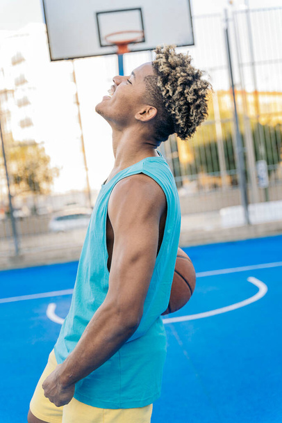 Happy afro american basketball player looking at camera holding the ball under his arm in a street basketball court. - Fotó, kép