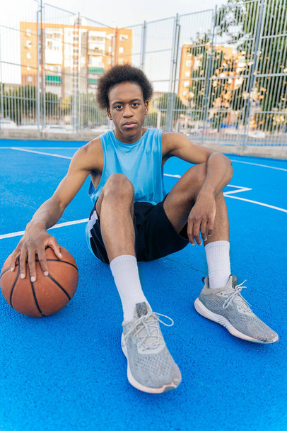 Portrait of an afro american basketball player crouching and holding the ball on the ground looking seriously at camera on the court. - Photo, image