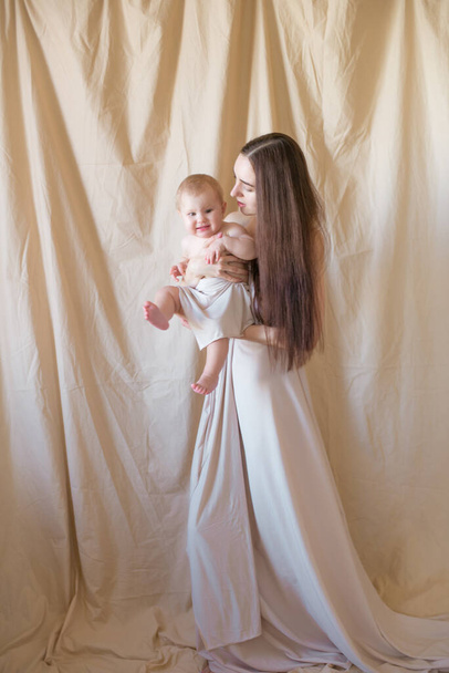 Young mother with long dark hair holding a cute little daughter in her arms in a nude fabric on a nude textile background. happy motherhood - Foto, immagini