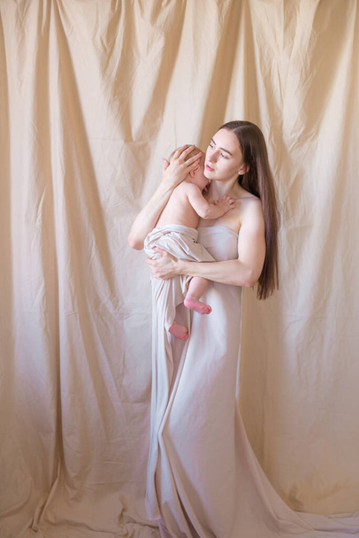 Young mother with long dark hair holding a cute little daughter in her arms in a nude fabric on a nude textile background. happy motherhood - Фото, изображение