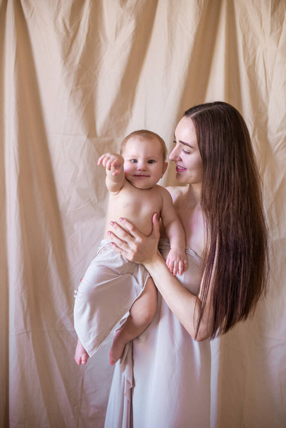 Young mother with long dark hair holding a cute little daughter in her arms in a nude fabric on a nude textile background. happy motherhood - Photo, image