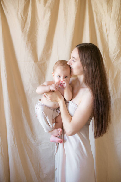 Young mother with long dark hair holding a cute little daughter in her arms in a nude fabric on a nude textile background. happy motherhood - Foto, Imagen