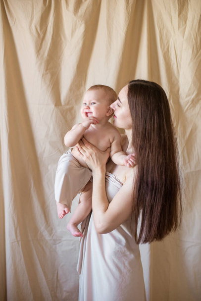 Young mother with long dark hair holding a cute little daughter in her arms in a nude fabric on a nude textile background. happy motherhood - Valokuva, kuva