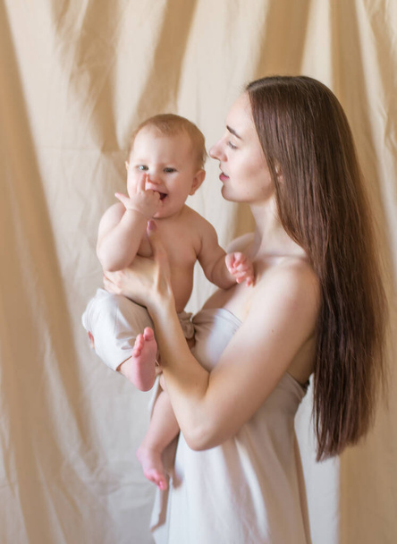 Young mother with long dark hair holding a cute little daughter in her arms in a nude fabric on a nude textile background. happy motherhood - Foto, Imagem