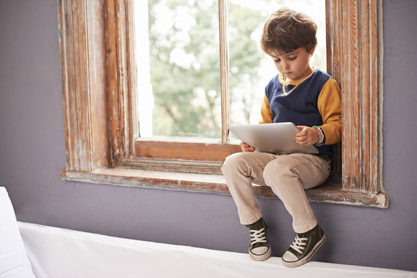 Hij is al technisch onderlegd. een schattig jongetje met behulp van een tablet tijdens het zitten op een vensterbank - Foto, afbeelding