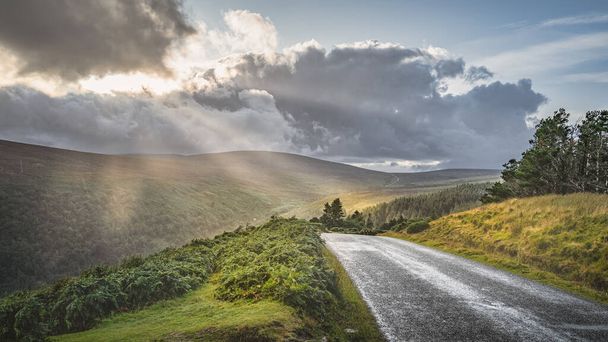 Carretera sinuosa que conduce a Lough Tay llamada The Guinness Lake iluminada por los rayos del sol desde el cielo dramático del atardecer con nubes de lluvia Wicklow Mountains, Irlanda - Foto, Imagen