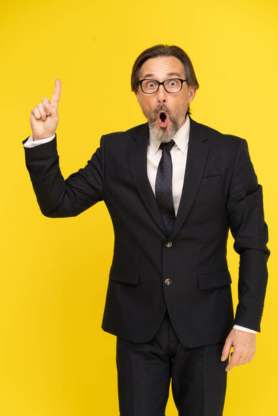 Shocked of offer pointing finger up middle aged businessman excited looking at camera isolated on yellow background. Handsome mature businessman in black suit and eye glasses. Business concept. - Fotografie, Obrázek