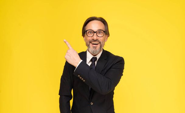 Mature grey beard businessman in black suit. Pointing sideways up middle aged business man in eye glasses smiling looking at camera isolated on yellow background. Business concept.  - Foto, Imagem