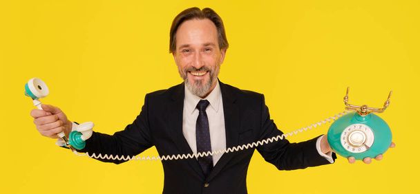 Middle aged handsome happy business man with old fashion telephone in hands stretched sideways looking at camera isolated on yellow background. Handsome businessman in black suit. Business concept.  - Foto, afbeelding