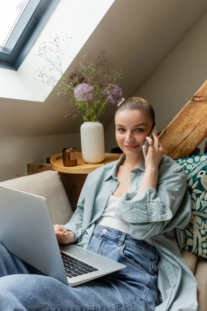 Smiling freelancer talking on smartphone and looking at camera near laptop at home  - Photo, Image