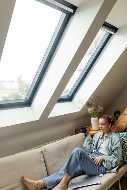 Jovem mulher no fone de ouvido usando smartphone e segurando copo no sofá em casa  - Foto, Imagem