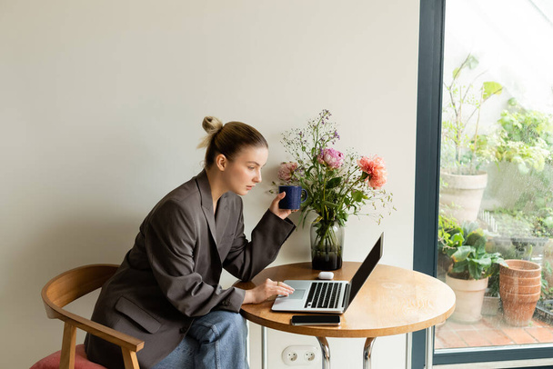Joven freelancer usando laptop y sosteniendo taza cerca de flores en casa  - Foto, Imagen