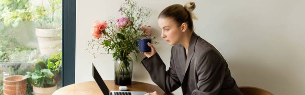 Donna in giacca in possesso di tazza e utilizzando computer portatile vicino a fiori a casa, banner  - Foto, immagini