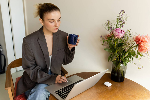 Young woman in blazer using laptop and holding cup near bouquet on table at home  - Foto, Imagen
