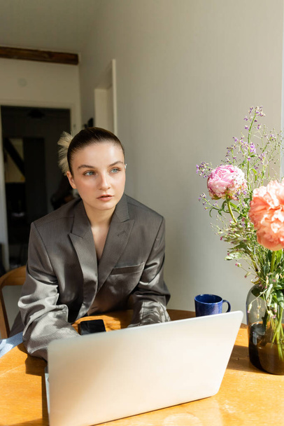 Young woman in blazer looking at flowers near devices and cup at home  - Fotografie, Obrázek