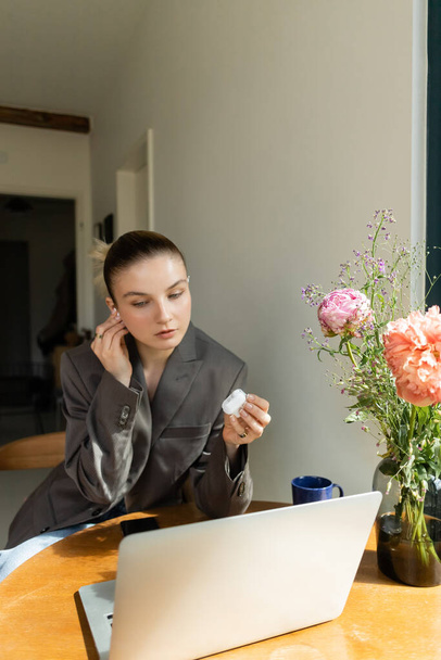 Kobieta w kurtce trzyma słuchawki w pobliżu urządzeń i kwiatów w domu  - Zdjęcie, obraz
