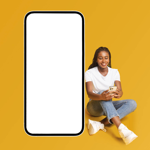Счастливая африканско-американская женщина с помощью телефона сидит на полу рядом с огромным смартфоном с пустым экраном изолированы на желтый, реклама отличная новая мобильная программа, веселая девушка представляет сайт - Фото, изображение