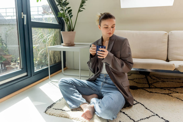 Woman in jacket holding cup while sitting on carpet in living room  - Foto, afbeelding