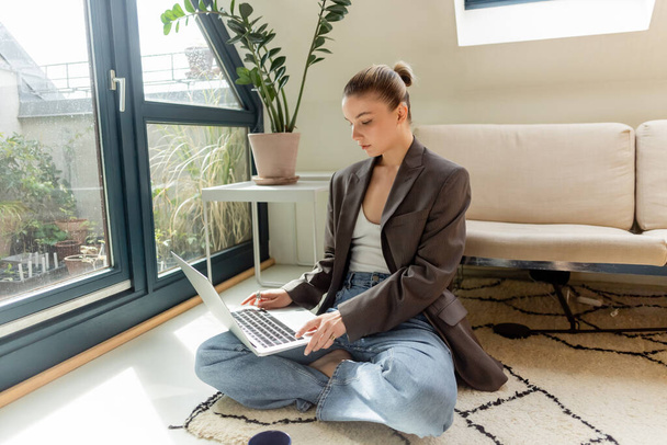 Freelancer in blazer using laptop while sitting on carpet at home  - Φωτογραφία, εικόνα