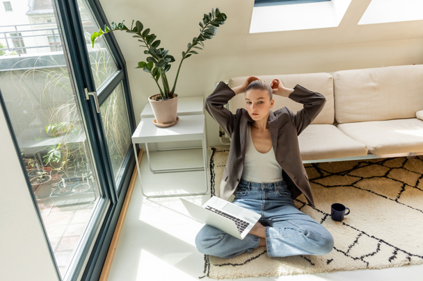 Freelancer in jacket touching hair near laptop and cup on floor at home  - Valokuva, kuva