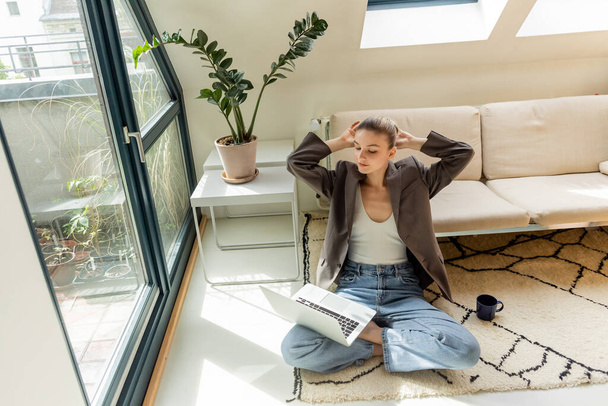 Femme en veste regardant ordinateur portable près de tasse sur le tapis à la maison  - Photo, image