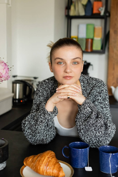 Young woman in knitted cardigan looking at camera near croissant and cups in kitchen  - Foto, Imagem
