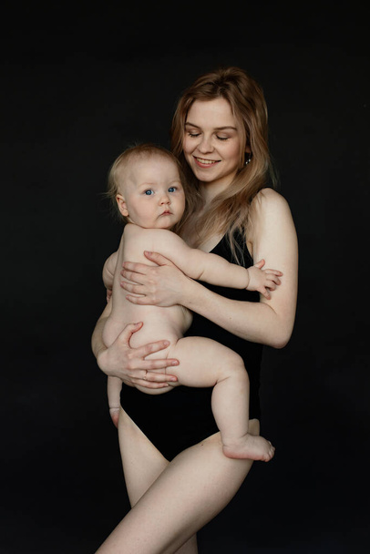Vertical portrait of blonde smiling woman, mother with close eyes holding and carrying her little baby in arms in black studio. Kid care, playing with child. Offspring development. Maternity vacation - Фото, зображення