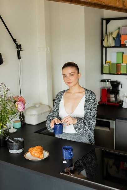 Young woman holding cup near croissant and flowers in kitchen  - Foto, Bild