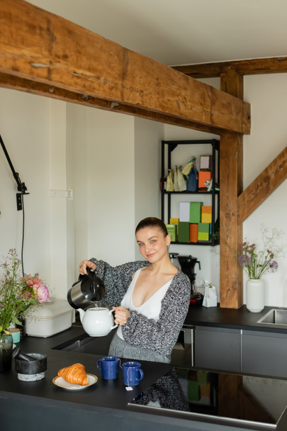 Smiling woman pouring water in teapot near cups and croissant in kitchen  - Fotoğraf, Görsel