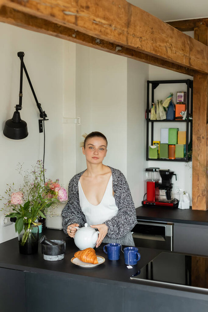 Young woman in knitted cardigan holding teapot near cups and croissant in kitchen  - Φωτογραφία, εικόνα