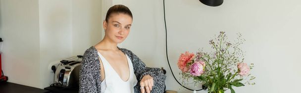Young woman in warm cardigan looking at camera near flowers in kitchen, banner  - Fotografie, Obrázek
