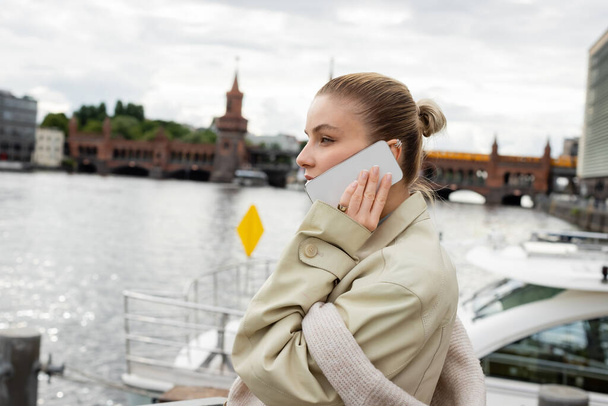 Widok z boku kobiety w płaszczu rozmawia przez telefon komórkowy w pobliżu rzeki w Berlinie  - Zdjęcie, obraz