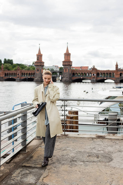 Jeune femme en trench coat tenant un sac d'embrayage et parlant sur smartphone à Berlin  - Photo, image