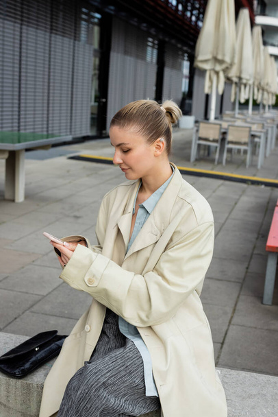 Jovem mulher em casaco de trincheira segurando celular perto de saco de embreagem ao ar livre em Berlim  - Foto, Imagem