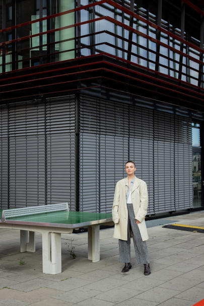Femme élégante en trench coat debout près de la table de ping-pong à l'extérieur à Berlin  - Photo, image
