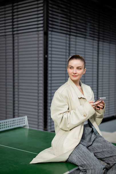 Woman in trench coat using smartphone while sitting on ping-pong table on street  - Φωτογραφία, εικόνα