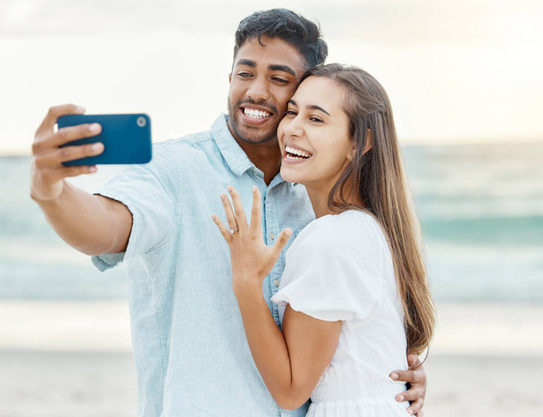 Любовь, пляж и пара после помолвки, делающая селфи на телефон во время летнего отпуска. Счастливая женщина показывает свое кольцо после предложения во время фотографирования с мужчиной во время отпуска - Фото, изображение