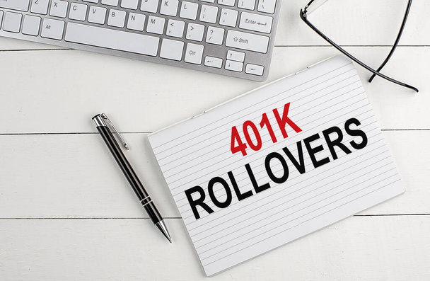 401K ROLLOVERS текст на ноутбуке с клавиатурой, ручки очки на белом деревянном фоне - Фото, изображение