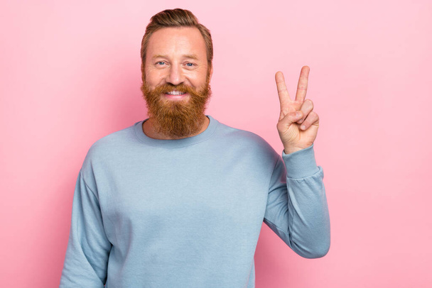 Portret van optimistisch vriendelijke man met rood kapsel baard dragen blauwe lange mouw tonen v-teken geïsoleerd op roze kleur achtergrond. - Foto, afbeelding