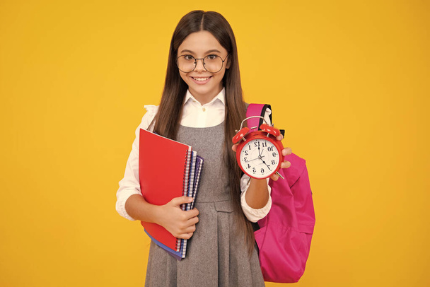 School teen girl checking time. Schoolgirl with clock alarm. School time. Happy face, positive and smiling emotions of teenager schoolgirl isolated on yellow - Photo, Image