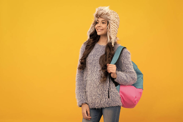 glückliches Kind mit lockigem Haar im Hut. zurück zur Schule. Teenie-Mädchen in Stricksachen auf gelbem Hintergrund. Kinder in warmer Kleidung tragen Rucksack. Ausdruck positiver Emotionen. Wintermode. - Foto, Bild